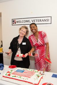 Two Clark staff members smiling and serving a U.S. flag-decorated cake under a sign that says "Welcome, Veterans!"