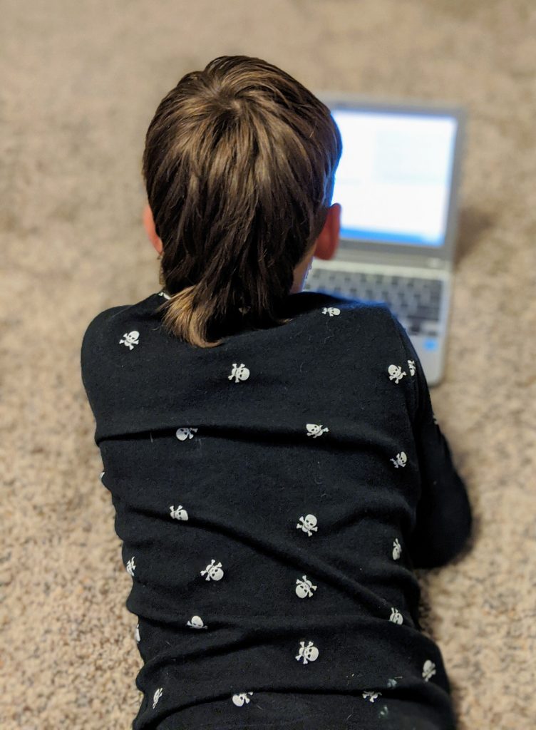 photo of 7-year-old child seen from the back, lying on floor and looking at laptop computer