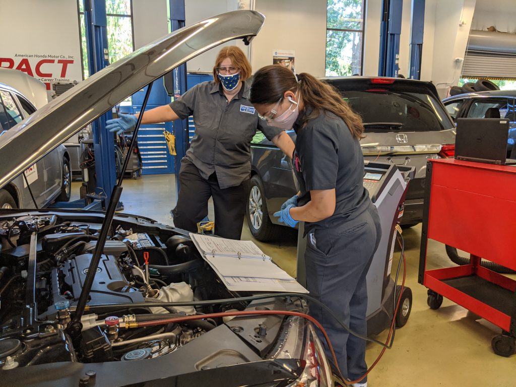 Two students looking at a car with an open hood, exposing the engine, and checking a notebook