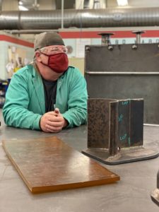 Welding student Jeff White, wearing cloth face mask and sitting in front of welded metal.