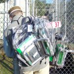 man seen from back, carrying multiple clear backpacks filled with folders, at a chain-link gate