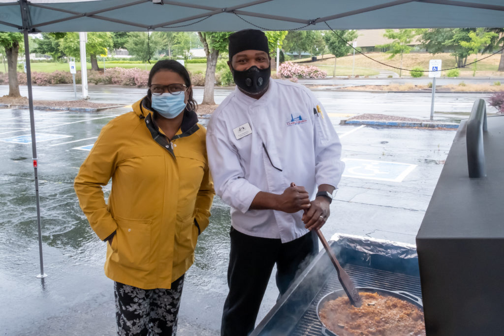 Dr. Karin Edwards in rainjacket and Chef Earl Frederick in chef's white jacket under a pop-up tent in the rain. Chef Earl is stirring some paella on a barbecue grill. Both are wearing face masks.