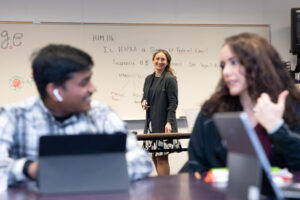 Professor Olga Lyubar stands at a whiteboard, smiling, while two students sit in front of her at tablet computers, talking to each other