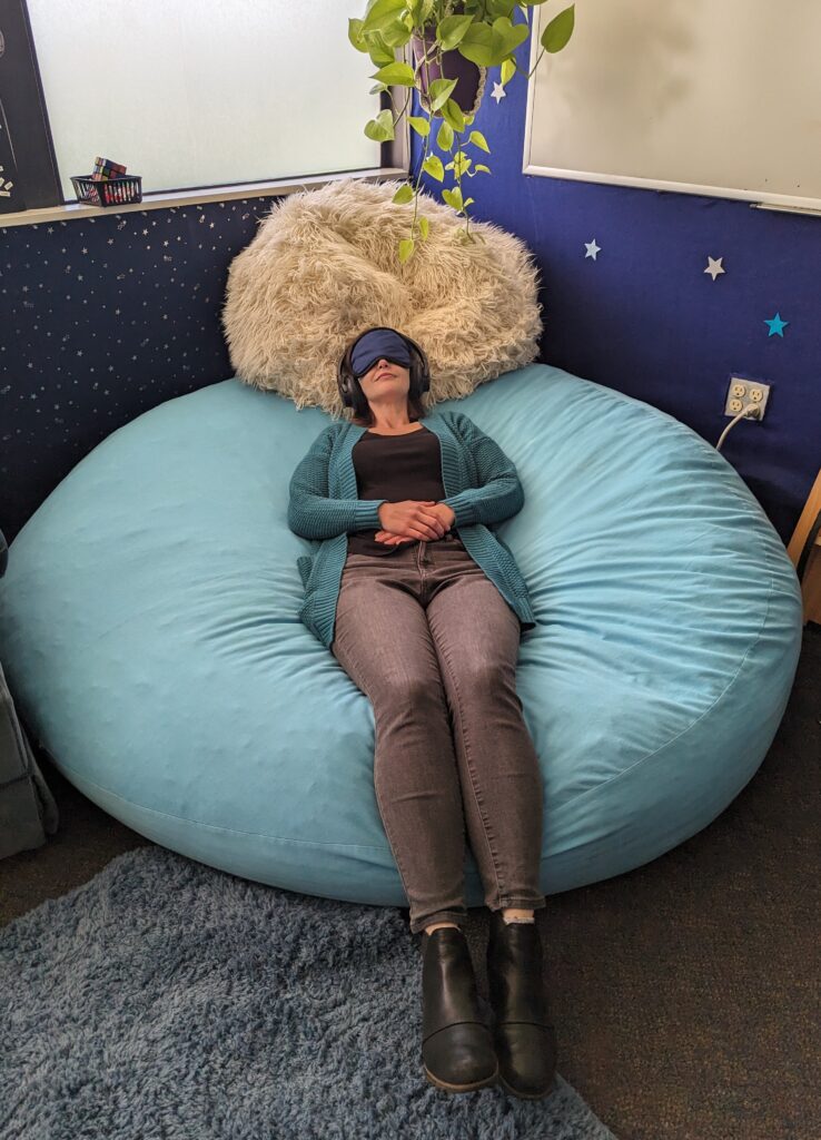 Counselor Shayna Collins demonstrates using noise-canceling headphones a mask and a beanbag to relax.