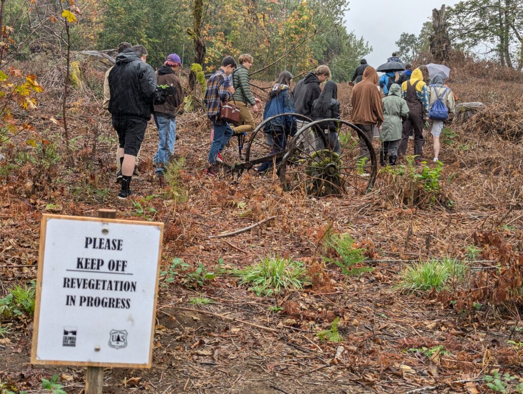 sign in front reads "please keep off. Revegetation in progress" people walk up a hill behind.