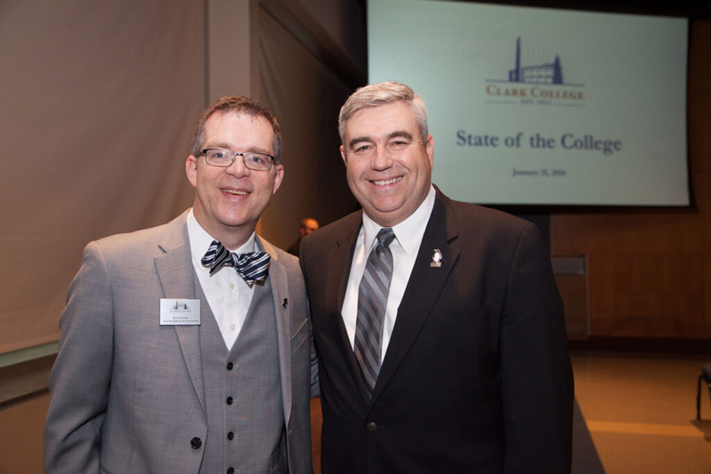 Two individuals wearing suits and smiling at the camera, with a screen behind them displaying the words, "State of the College".