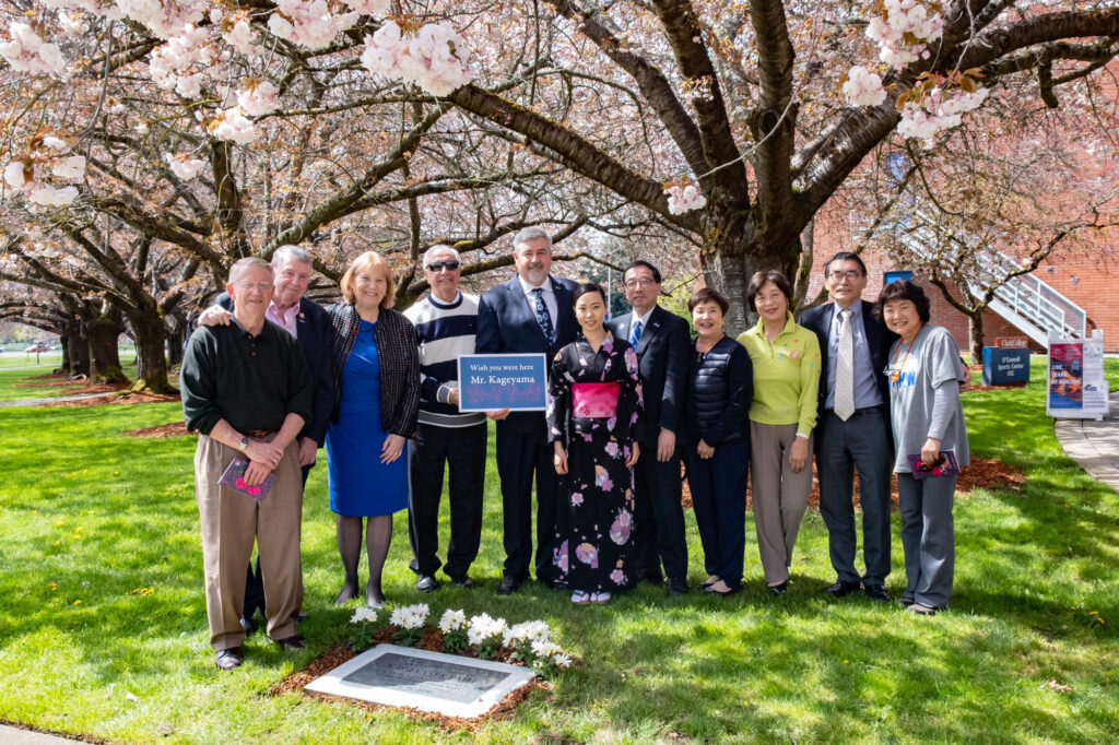 A group of people, including the former Clark College president holding a sign that reads "Wish you were here, Mr. Kageyama". They are standing in front of a stone plaque that reads "Kageyama Path"