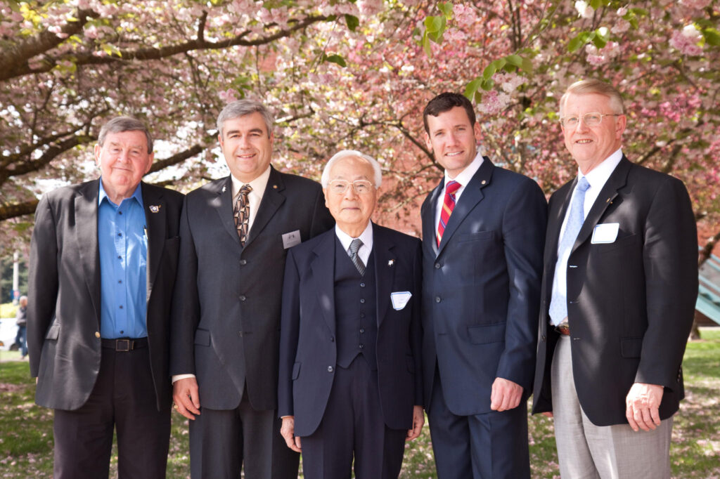 A group of individuals standing together with a row of cherry blossom trees in the background.