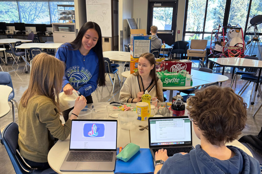 students sit at a table with laptops and are mixing liquids in containers.