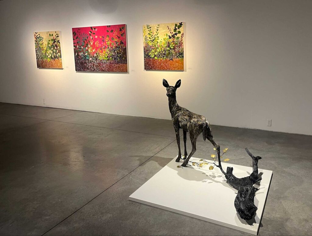 An art exhibition featuring three paintings on a wall and a statue of a deer with a log on display.