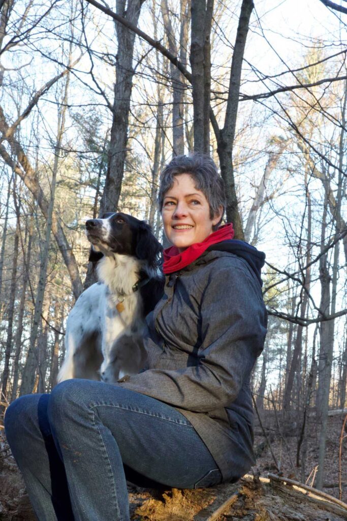 A woman sitting in a forest with her dog, smiling and looking away from the camera.