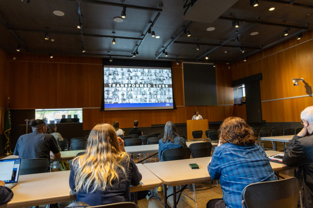 people watch talk with large screen that reads "transformed lives"