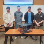 four people stand behind a table with a transparent box