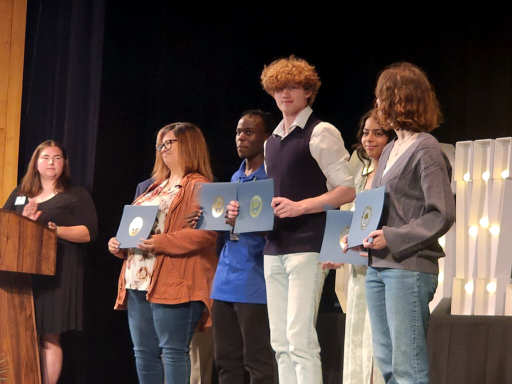 Person clapping for 5 people standing on stage with award certificates.