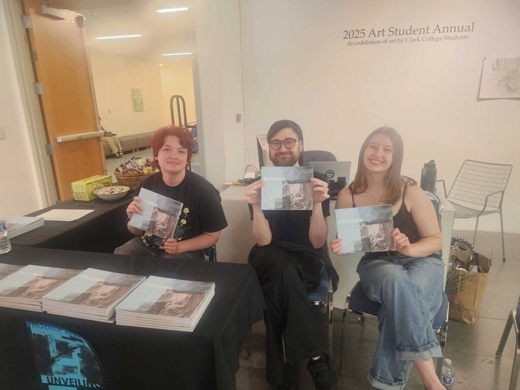 Three people sitting at a table with books, smiling at the camera while holding a copy of The Phoenix.