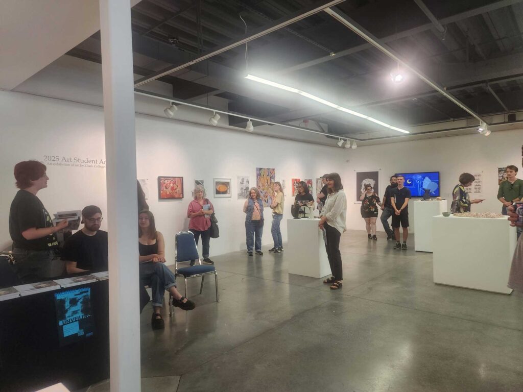A group of art gallery attendees gathered and attentively listening to a speaker presenting in the corner of the room.