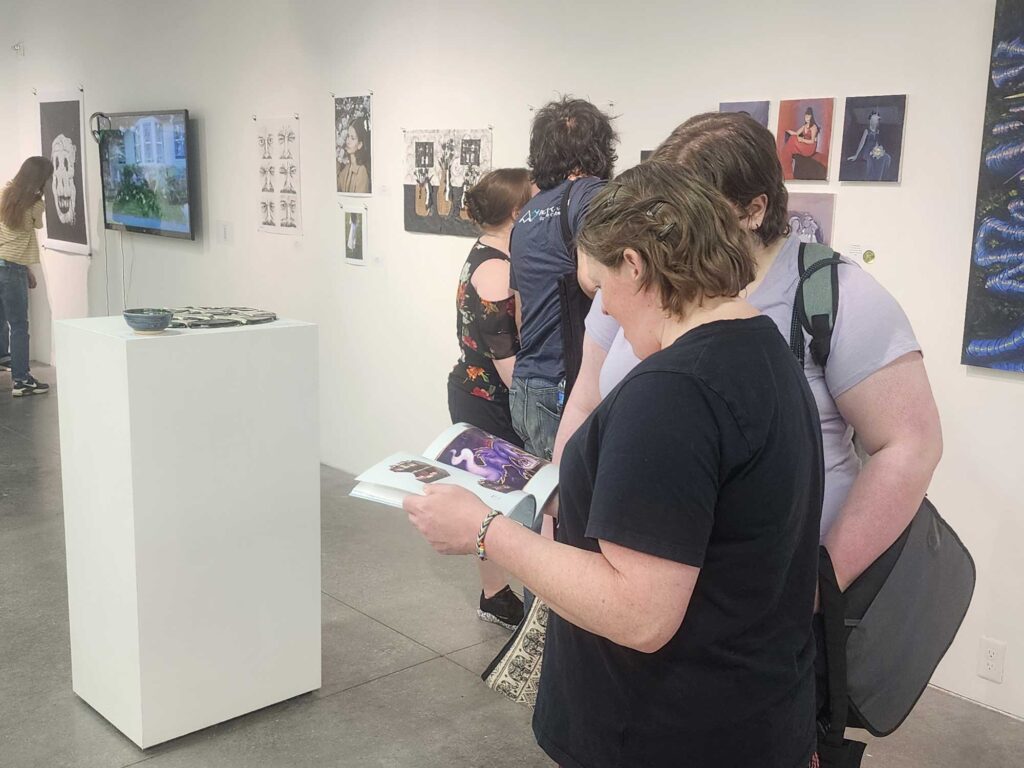 Two people looking at a brochure with pictures inside an art gallery, with other attendees in the background viewing artwork on the walls.
