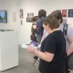 Two people looking at a brochure with pictures inside an art gallery, with other attendees in the background viewing artwork on the walls.