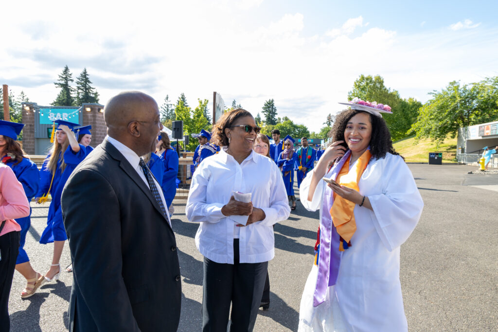 three people stand outside one wearing a white cap and gown