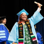 student holds scroll above their head wearing cap and gown with stole that reads 2025