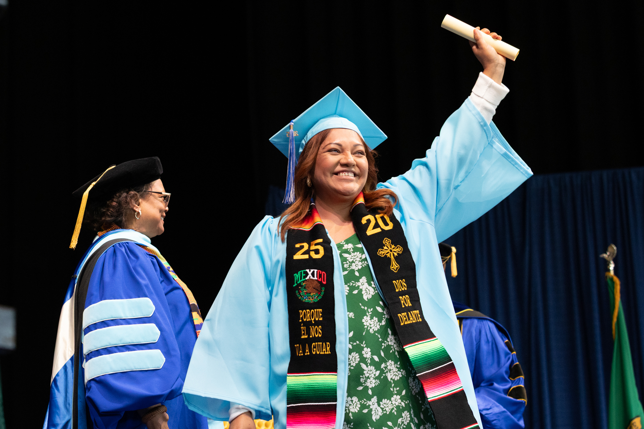 student holds scroll above their head wearing cap and gown with stole that reads 2025
