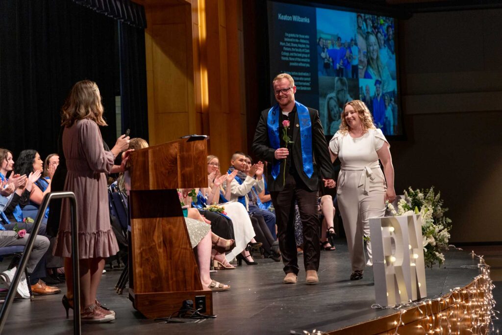 A nursing graduate walking down the stage with his wife, smiling and holding a single rose.