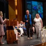 A nursing graduate walking down the stage with a companion, smiling and holding a single rose.