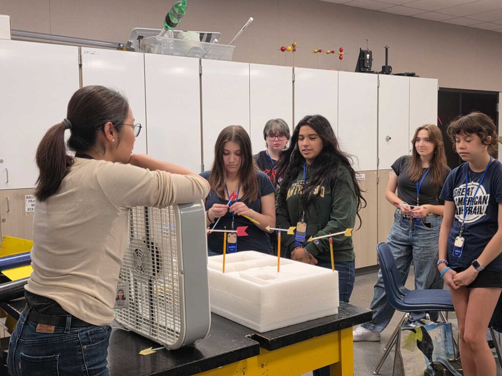 A professor leaning on a fan, speaking to a group of students while demonstrating with a styrofoam model in front of them, featuring pencils and arrows taped on top to illustrate concepts like center of gravity and flight stability.