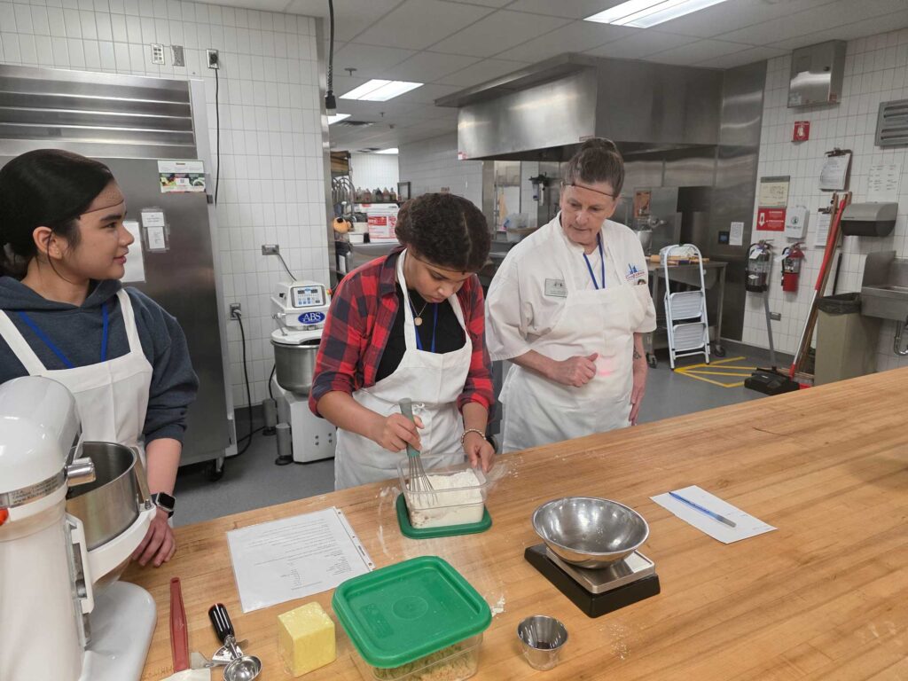 A chef and student wearing aprons and hairnets; the chef is guiding the student who is whipping something in a container.