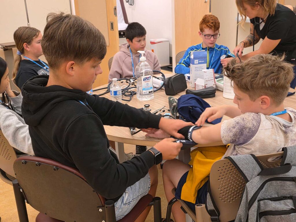 Students gathered around a table learning how to measure blood pressure, with two kids in focus practicing by putting on the arm cuff.
