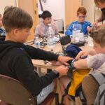Students gathered around a table learning how to measure blood pressure, with two kids in focus practicing by putting on the arm cuff.