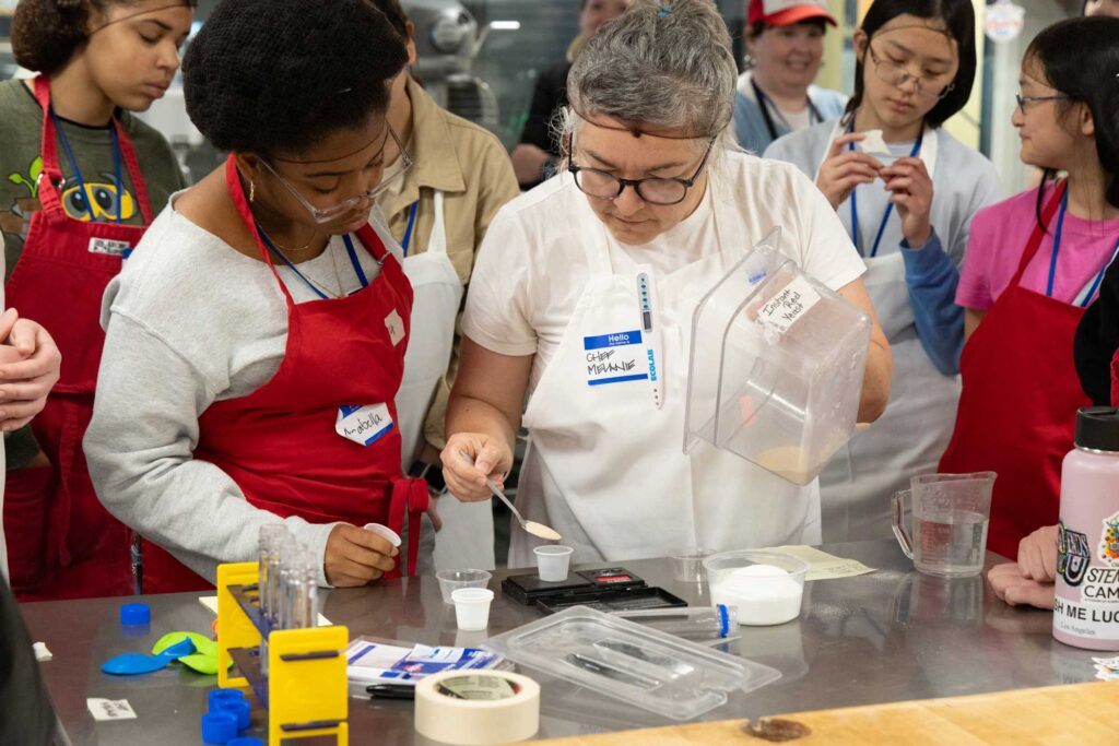 A chef scooping instant red yeast into a small plastic cup on a measuring scale, with a student beside her watching attentively.