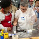 A chef scooping instant red yeast into a small plastic cup on a measuring scale, with a student beside her watching attentively.