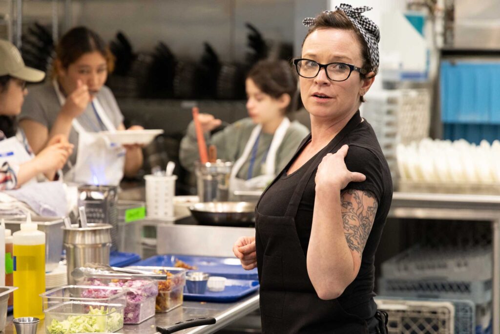 A chef turning to look behind her while students in the background perform a taste test.