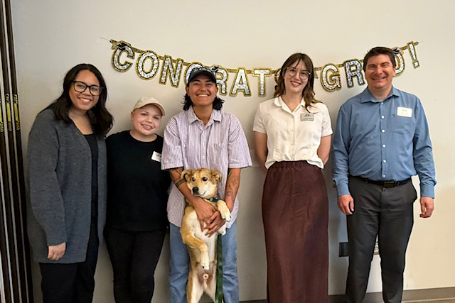 Five people stand with a dog and with sign that reads "congrats grads" behind them.