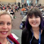 two people smile at camera with people at desks behind them