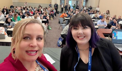two people smile at camera with people at desks behind them